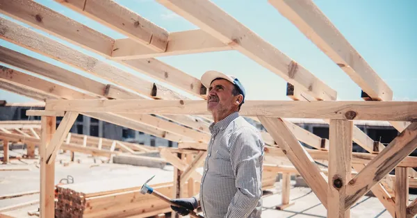 A construction worker surveys a wooden roof frame build on a sunny day. construction worker inspecting roof frame outdoors
