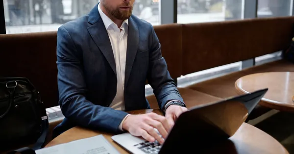 A professional businessman in a suit working on a laptop inside a cafe setting, focusing on a report. man wearing suit jacket using a laptop