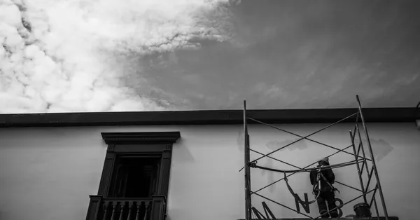 A worker paints a building wall on scaffolding under a cloudy sky. Black and white. Urban scene. worker on scaffolding painting building exterior
