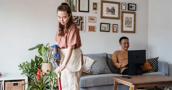 An Asian woman vacuuming while a man works on a laptop in a modern living room. a woman vacuuming in a living room