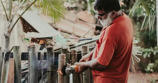 An adult man wearing a red shirt uses a grinder on a wooden fence with sparks flying, showcasing outdoor work. man in red shirt holding a power tool