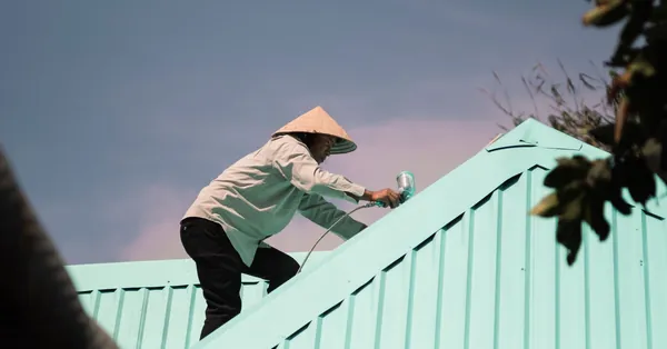 Man in conical hat paints a rooftop, engaging in outdoor work. Low angle view. man in conical hat working on roof
