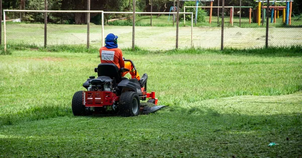 Individual operating a riding lawnmower on a grassy field in a recreational park, Londrina, Brazil. person driving a lawnmower
