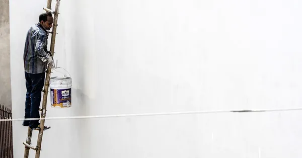 A worker on a ladder paints a large white wall outdoors, showcasing construction work. worker painting wall on ladder in urban setting