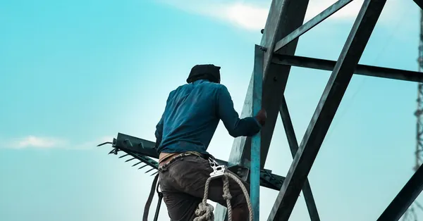 A construction worker climbs a steel tower with safety gear against a clear sky in Mymensingh, Bangladesh. construction worker climbing high steel structure